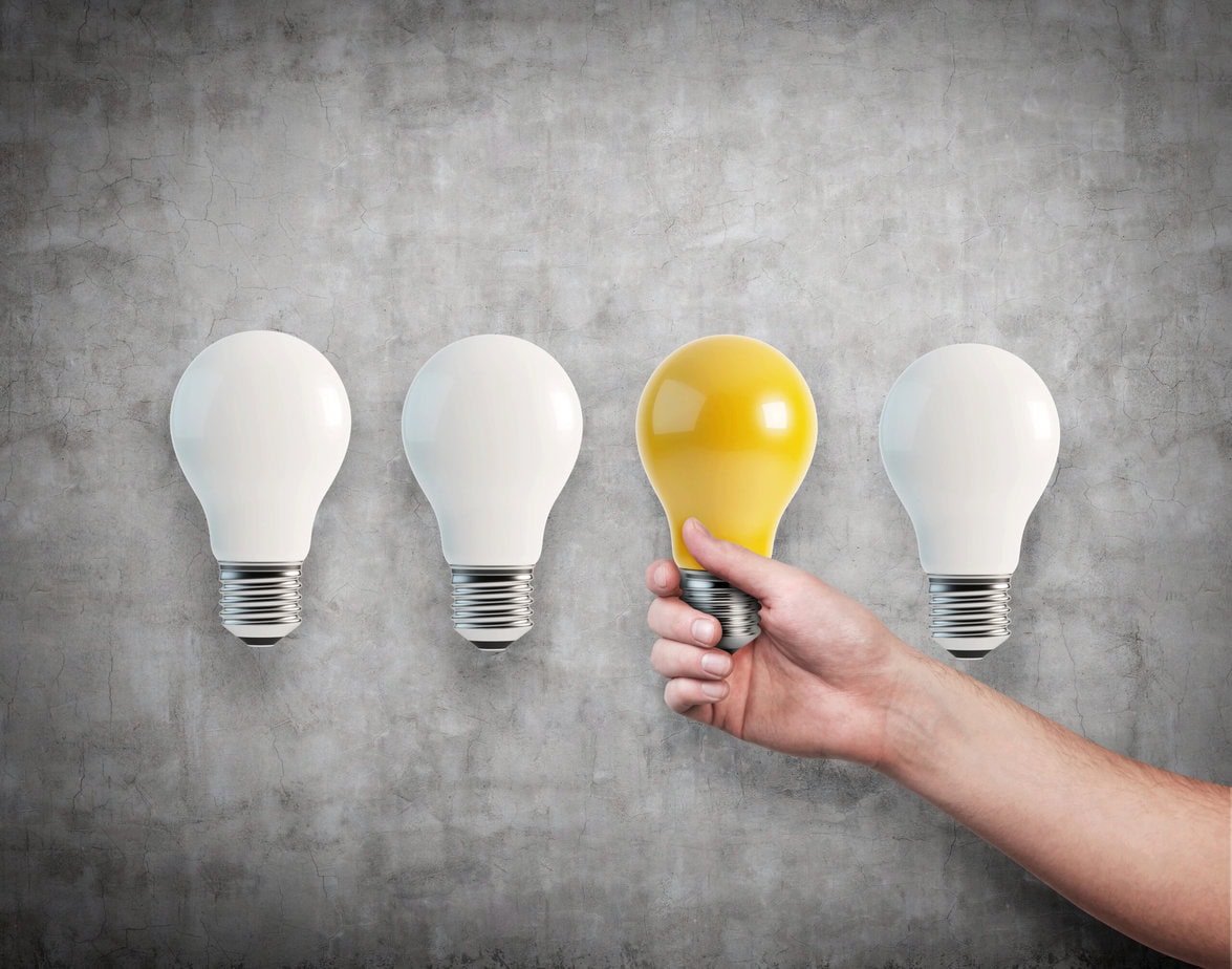 A hand holds a yellow light bulb next to three unlit white bulbs against a gray textured wall.